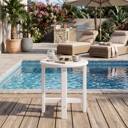 A small round white outdoor bar table with legs, shown on a wooden deck beside a swimming pool. The table is styled with a glass, a book, and a small cup on top.