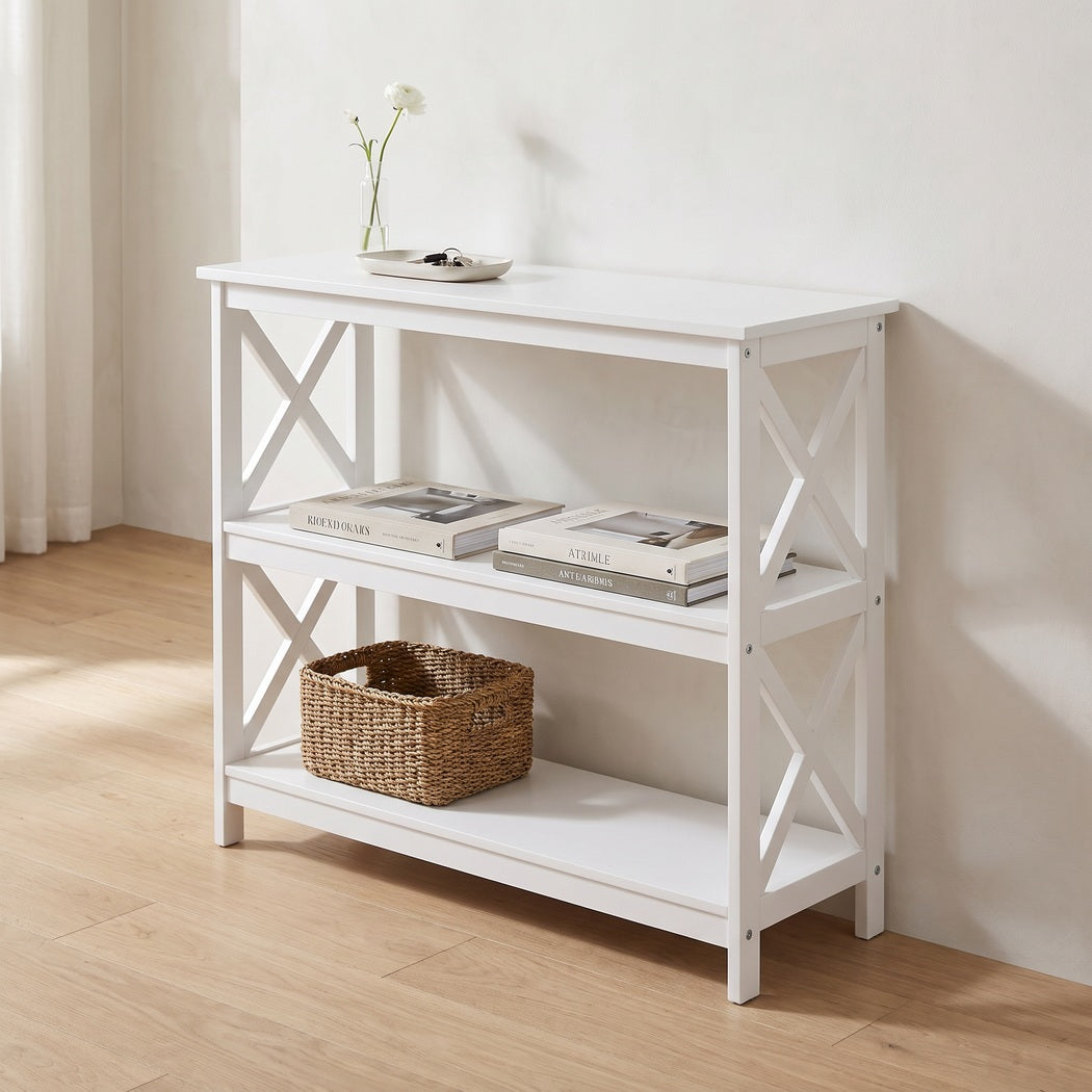White three-tier console table against a plaster hallway wall with a key tray, books and a woven basket