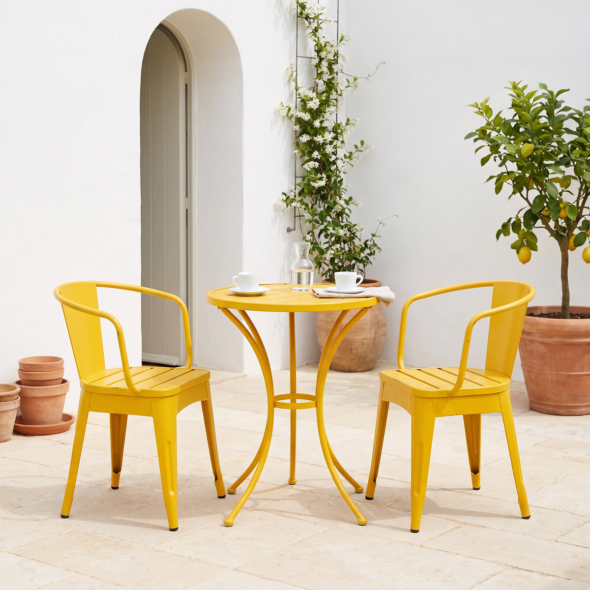 Yellow bistro set with two cups and a carafe on a sunlit courtyard terrace beside an arched doorway and a lemon tree